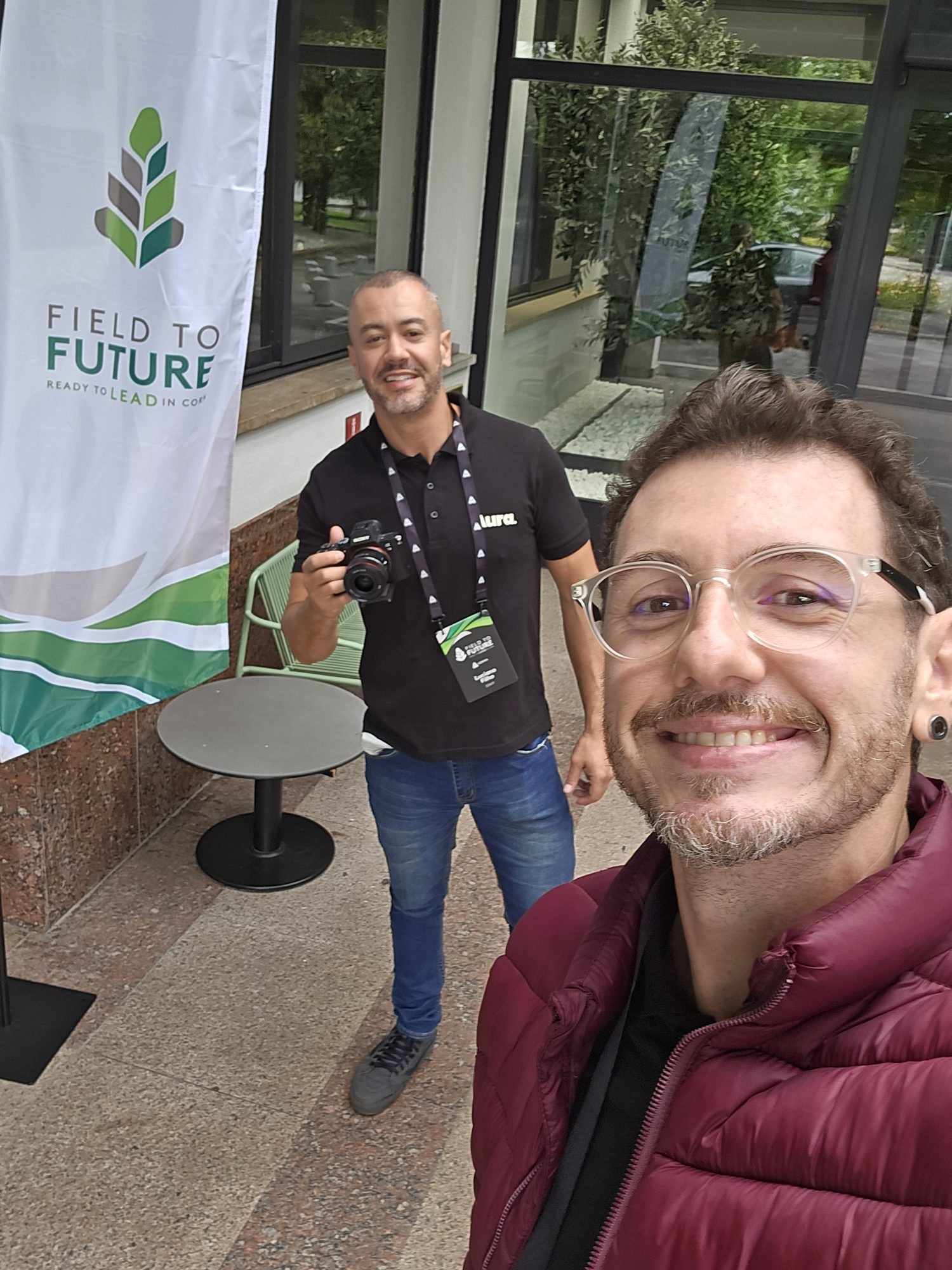 Field to Future, Milão 2026: selfie de dois integrantes da equipe diante de uma bandeira Field to Future