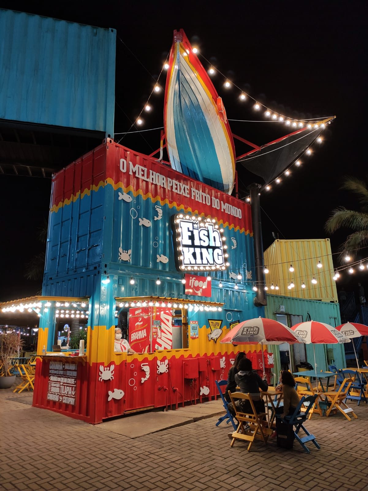 Fachada em container iluminada à noite, com mesas no parque
